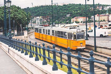 Fototapeta premium Budapest setting of vintage yellow tram traveling along the river embankment tracks. Decorative metal fence frames the foreground while lush green hills and a bridge stand in urban distance