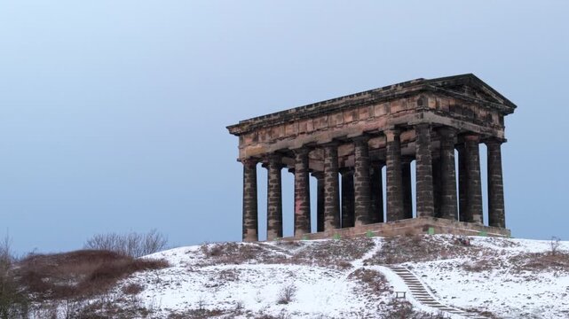 Low-angle telephoto drone shot rising behind the snow-covered hill to reveal the Penshaw Monument in Sunderland against a winter sky.