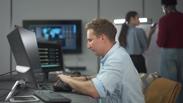 Young male software engineer writing program code on multiple desktop screens while his colleagues working in the background in a modern office.