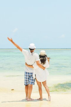 Couple stand together hugging hug enjoy arms up on white sand beach in sunny tropical destination. Romantic summer holiday by turquoise ocean with clear blue sky. Honeymoon paradise vibes
