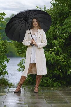 Woman in light raincoat and brown boots stands with black umbrella near river in park in rain. Trees and plants surround her. Rain creates wet path and cloudy sky above
