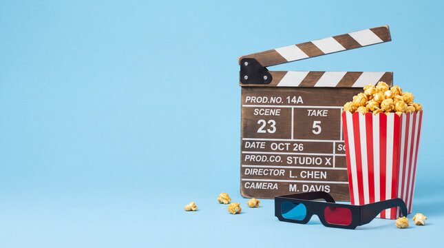 A movie clapperboard, a striped bucket of popcorn, and 3D glasses arranged together on a solid blue background, ideal for cinema and film concepts.