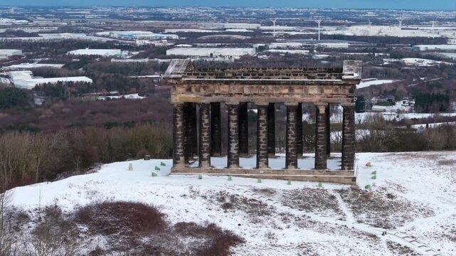 Aerial drone orbit shot of the snow-covered Penshaw Monument in Sunderland, showcasing the Nissan car plant and wind turbines across the winter landscape in the background.