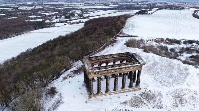 Elevated high-angle aerial drone orbit looking down on the snow-covered Penshaw Monument on Penshaw Hill in Sunderland, surrounded by winter countryside