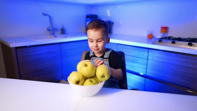 Boy reaching for yellow apples in kitchen. A white fruit bowl fills the foreground under vivid blue indoor lighting.