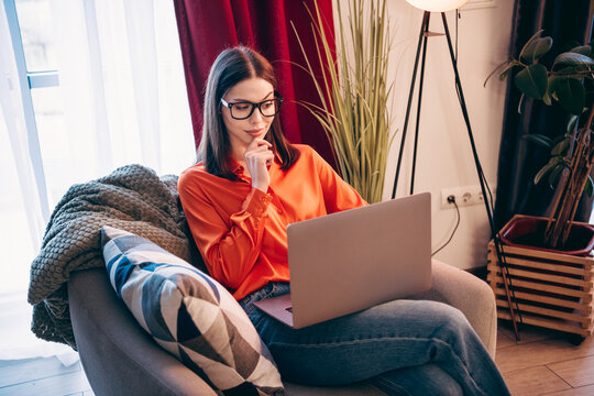Young woman working on a laptop at home, remote work and study in a cozy living room with thoughtful expression