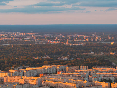 Aerial photograph of long rows of white prefabricated apartment blocks, courtyards and access roads in Ziepniekkalns, a large residential district of Riga, in warm golden hour light.