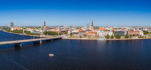 Naklejka premium Panoramic aerial drone view of the UNESCO-listed centre of Riga rising behind the wide Daugava river, its quays, bridges and church spires seen from the air, under a clear blue summer sky.