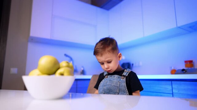 Boy looking down beside bowl of yellow apples. A wide composition shows the seated child and glossy tabletop under blue light.