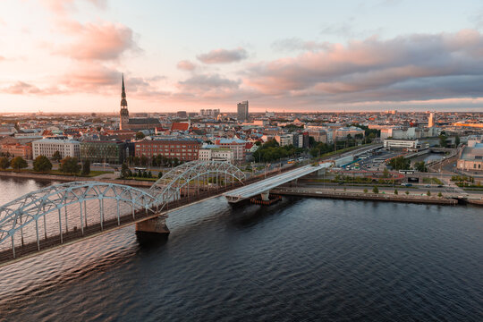 Aerial drone view of the riveted steel arches of the Riga railway bridge spanning the wide Daugava river towards the city centre, at sunset.