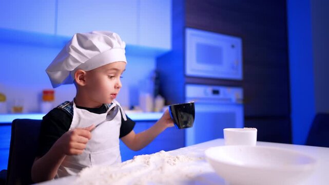 Boy holding spoon and mug beside flour. Photographed in profile, the child glances downward near a microwave oven in soft blue light.