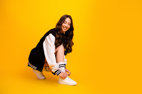 Smiling young woman tying sneakers in varsity jacket on bright yellow background