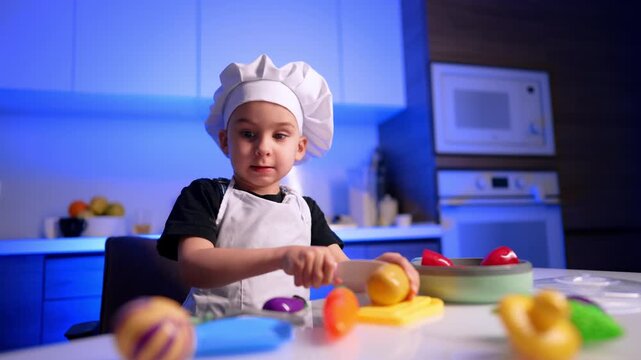 Boy cutting toy lemon on yellow board. Fresh fruit rests in a bowl behind him while a microwave oven appears blurred in the background.