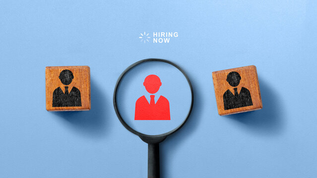 Flat lay of a magnifying glass highlighting a red candidate icon between wooden blocks on a blue background with 'Hiring Now' text. HR Recruitment concept