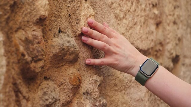 Close-up of a woman's hand gently touching the rough surface of an ancient stone wall while walking through a historic European street. Travel, heritage, architecture and tourism concept. High quality