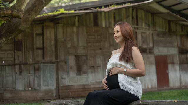 Smiling pregnant woman in a white top holding her belly while sitting outdoors in front of a weathered rustic wooden house.