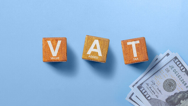 Wooden blocks spelling VAT (Value Added Tax) with US dollar bills on a blue background, symbolizing business finance and taxation. Financial regulation and tax compliance concept