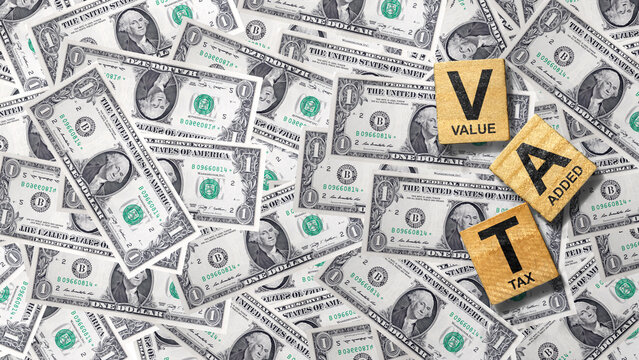 Wooden blocks spelling VAT (Value Added Tax) on a background of scattered US dollar bills, representing finance and taxation. Financial regulation and tax compliance concept