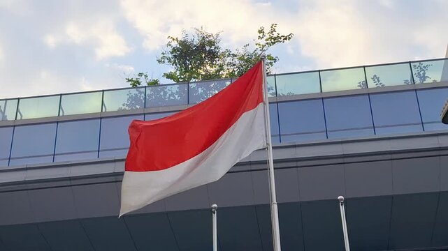 Red and white flag waves in front of modern glass building. Sky shows soft clouds above the structure. Flagpole stands tall beside sleek architectural lines