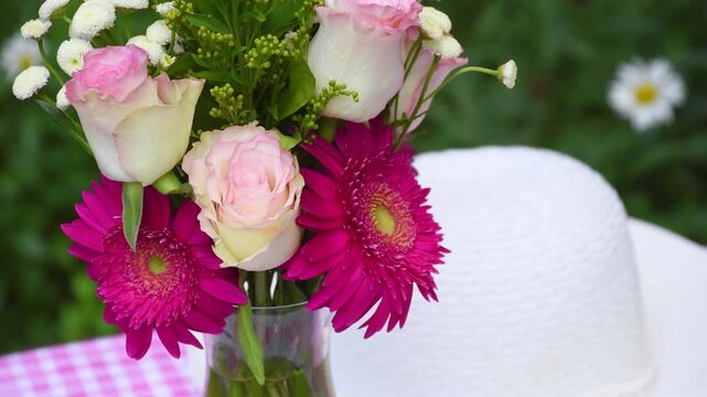 Elegant pink floral arrangement displayed with a white sun hat on a pink and white gingham table cloth with green foliage in the background. Pink roses and fuchsia gerbera daisy flowers in a vase