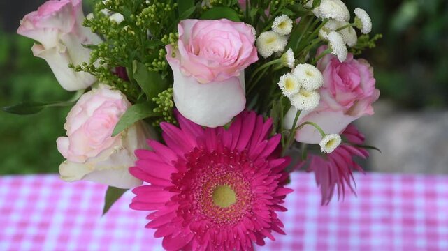 Elegant pink floral arrangement displayed on a pink and white gingham table cloth with green foliage in the background. Pink roses and fuchsia Gerbera daisy flowers in a vase