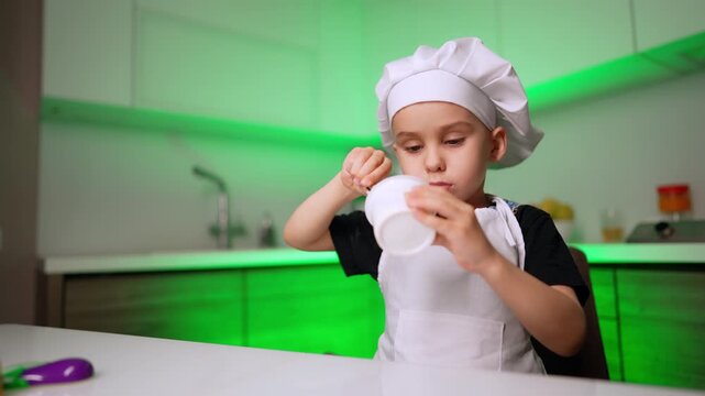 Little boy in chef hat drinking from plastic cup. A young child wearing an apron tilts a white container to eat the remaining food in a green backlit kitchen.