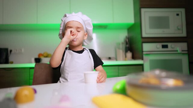 Little boy in chef hat tasting food in kitchen. A young child wearing a white apron licks yogurt from his face while holding a plastic cup at a white table.