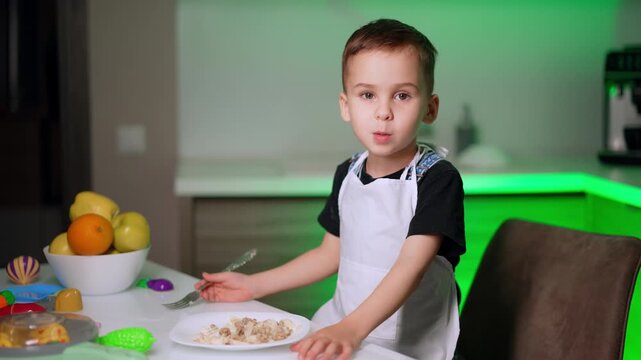 Young boy in apron holding fork and looking at camera. A child sits at a white table with a plate of food and a bowl of fruit under green lighting in a modern kitchen.