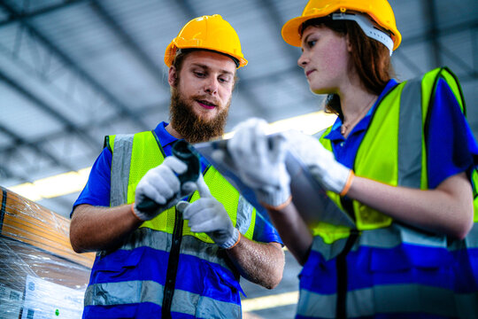 Bearded male warehouse worker and young female colleague in hard hats and safety vests scan carton barcodes with a handheld reader in a large fulfilment centre with rows of stacked pallets.