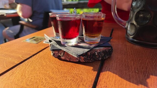 Sunlit outdoor table scene shows three clear glasses filled with amber and ruby drinks resting on rough bark coaster, with ice, soft reflections, rustic wood grain, and blurred guests behind
