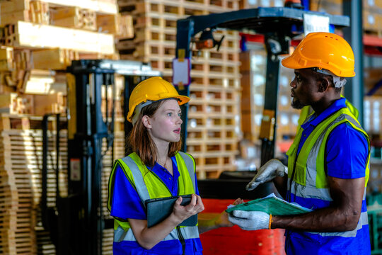 Black male logistics employee and his female coworker wearing hard hats and hi-vis vests resolve a stock question in a quick conversation in a bright modern distribution warehouse with tall.