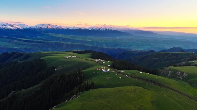 Aerial sunset over green alpine grasslands with nomadic yurt camps and snow-capped mountains in Kalajun Grassland, Xinjiang, China.