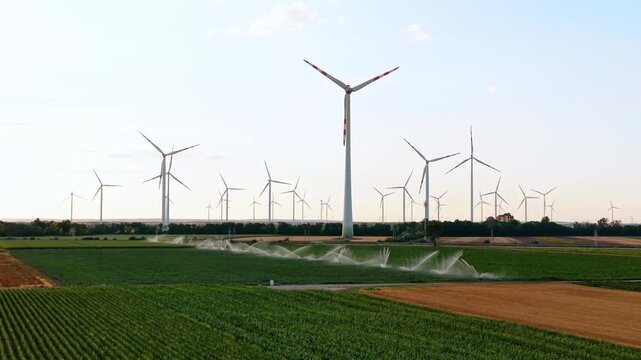 Wind turbine towers above irrigated cropland. Numerous turbines line the horizon while sprinklers water a patchwork of green and brown fields below.