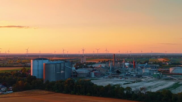Industrial facility silhouetted against wind farm sunset. Grain silos and a tall chimney stand before rows of turbines under an orange and pink evening sky.