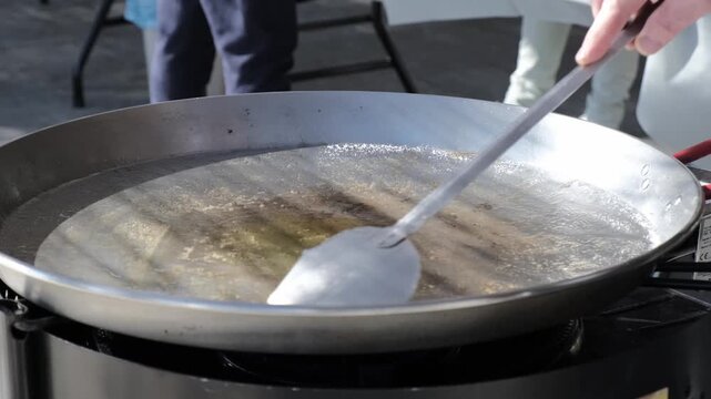 A large metal frying pan and a slotted spoon during outdoor cooking; in the background, blurred figures of people busy cooking. Streetfood festival