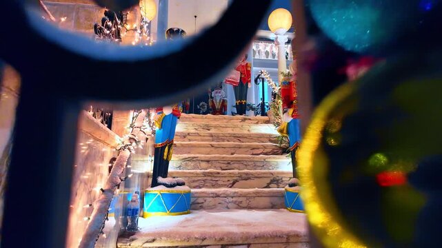 Nutcracker statues on snow covered marble steps. White holiday lights and decorative figures line the outdoor staircase viewed through a metal fence.