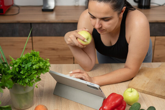 A Caucasian woman at home in the kitchen eats an apple and looks at a vegetable smoothie recipe on her tablet, the concept of Detcox and fresh vitamins
