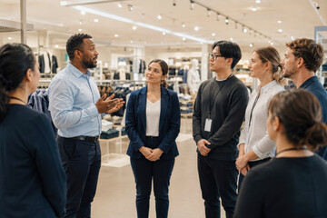 Obraz premium Retail team gathers listening to manager giving instructions in a clothing store. Employees stand in a circle focused and engaged during a briefing.