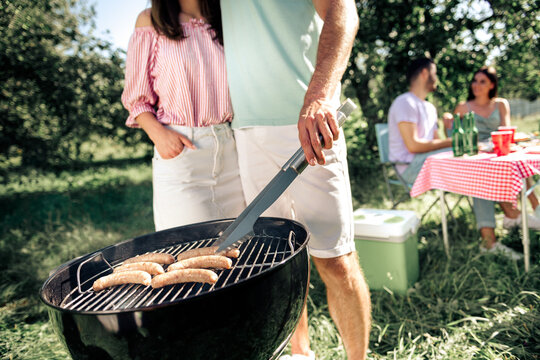 Couple grilling sausages on a charcoal barbecue at a summer picnic with friends in a sunny garden