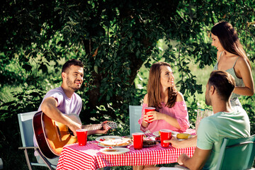 Fototapeta premium Friends enjoying a summer picnic outdoors with guitar, food and red cups at a checkered tablecloth under trees