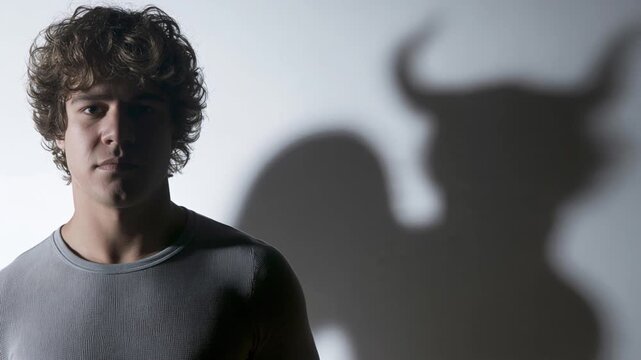 Young man looking at camera with devil horned shadow on wall behind him. Inner demon, dark side and psychological alter ego concept