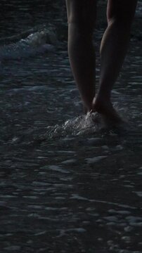 Barefoot hiker traverses tide-swept shoreline at twilight, crossing dark wet sand while cool waves flow over skin. Static vertical medium shot.