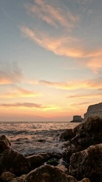 Ocean waves crash against dark rocks during a serene sunset on the horizon. Fixed vertical angle shot.
