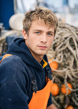 Young fisherman looks into camera while wearing orange overalls and navy hoodie near fishing nets and buoys on a dock. Natural expression and maritime workwear convey hardworking mood.