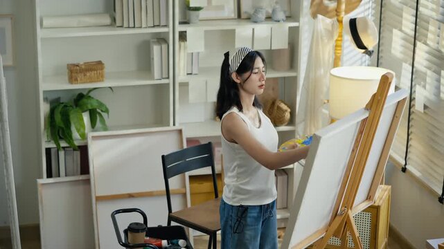 Young Asian woman artist painting on canvas easel with paintbrush in art studio, representing creative professional women designer, fine arts student lifestyle and freelance illustrator