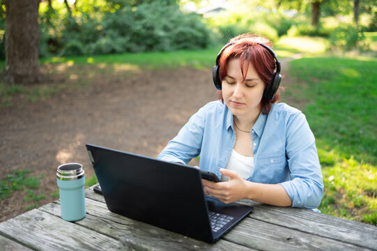 Young woman working on laptop at wooden table in summer park. Outdoor remote office concept.