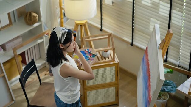 Young Asian woman artist painting on canvas easel with paintbrush in art studio, representing creative professional women designer, fine arts student lifestyle and freelance illustrator
