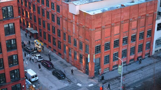 Red brick warehouses along DUMBO street. Pedestrians, parked cars and a food cart occupy the cobbled intersection below.