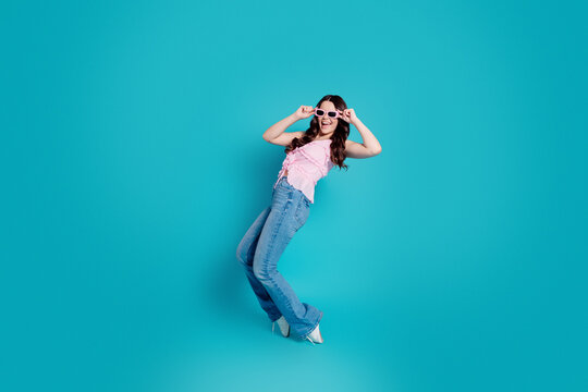 Young woman in pink top and jeans wearing sunglasses and smiling against a vibrant turquoise studio background &mdash; playful fashion portrait