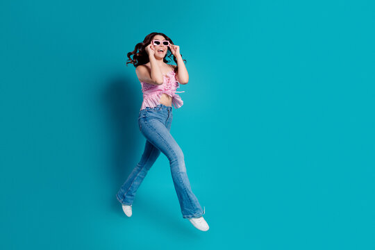 Happy young woman jumping in pink top and jeans wearing sunglasses on turquoise background, summer fashion lifestyle and joyful portrait